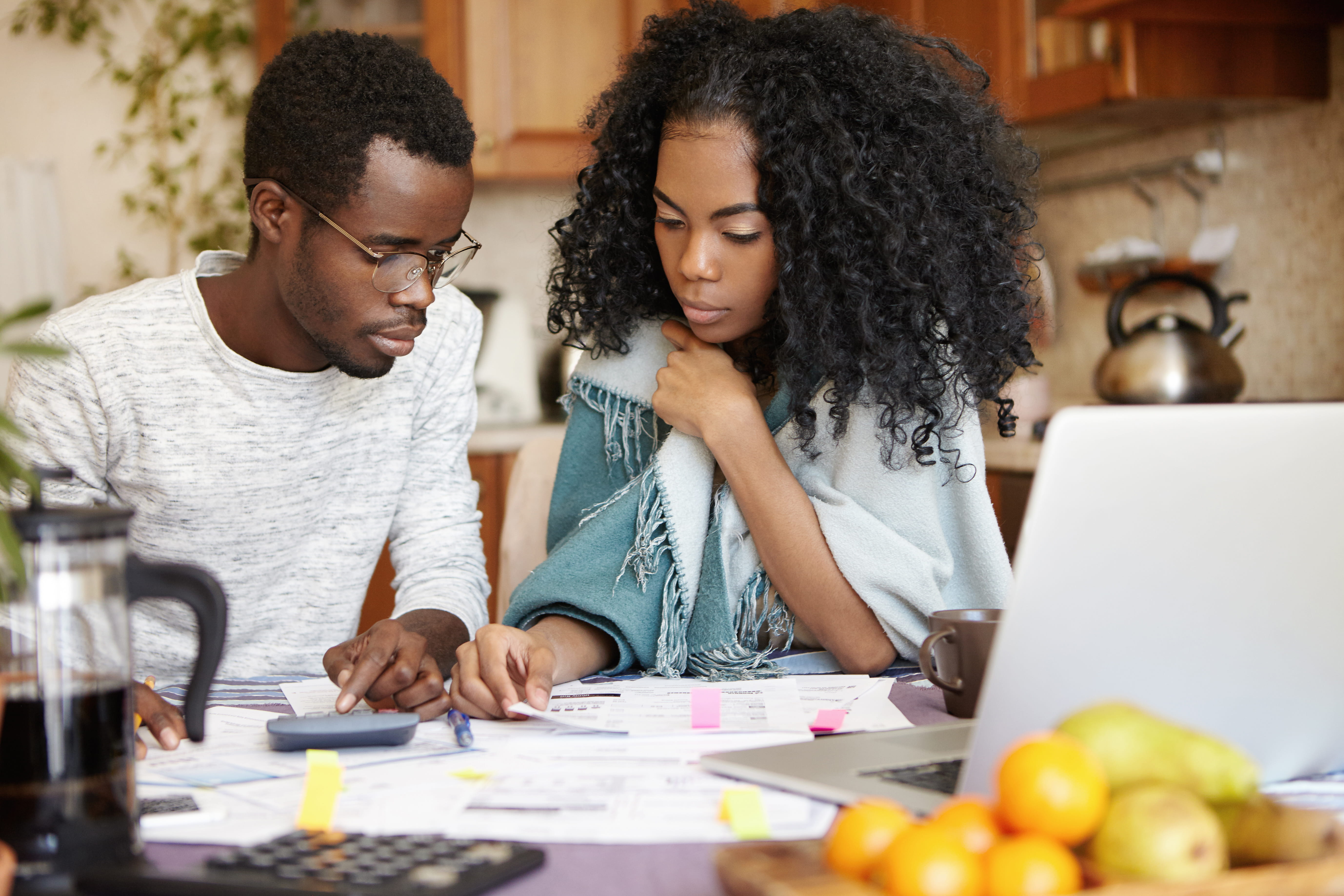 A Black couple focuses intently on financial paperwork and a calculator at a kitchen table with a laptop.