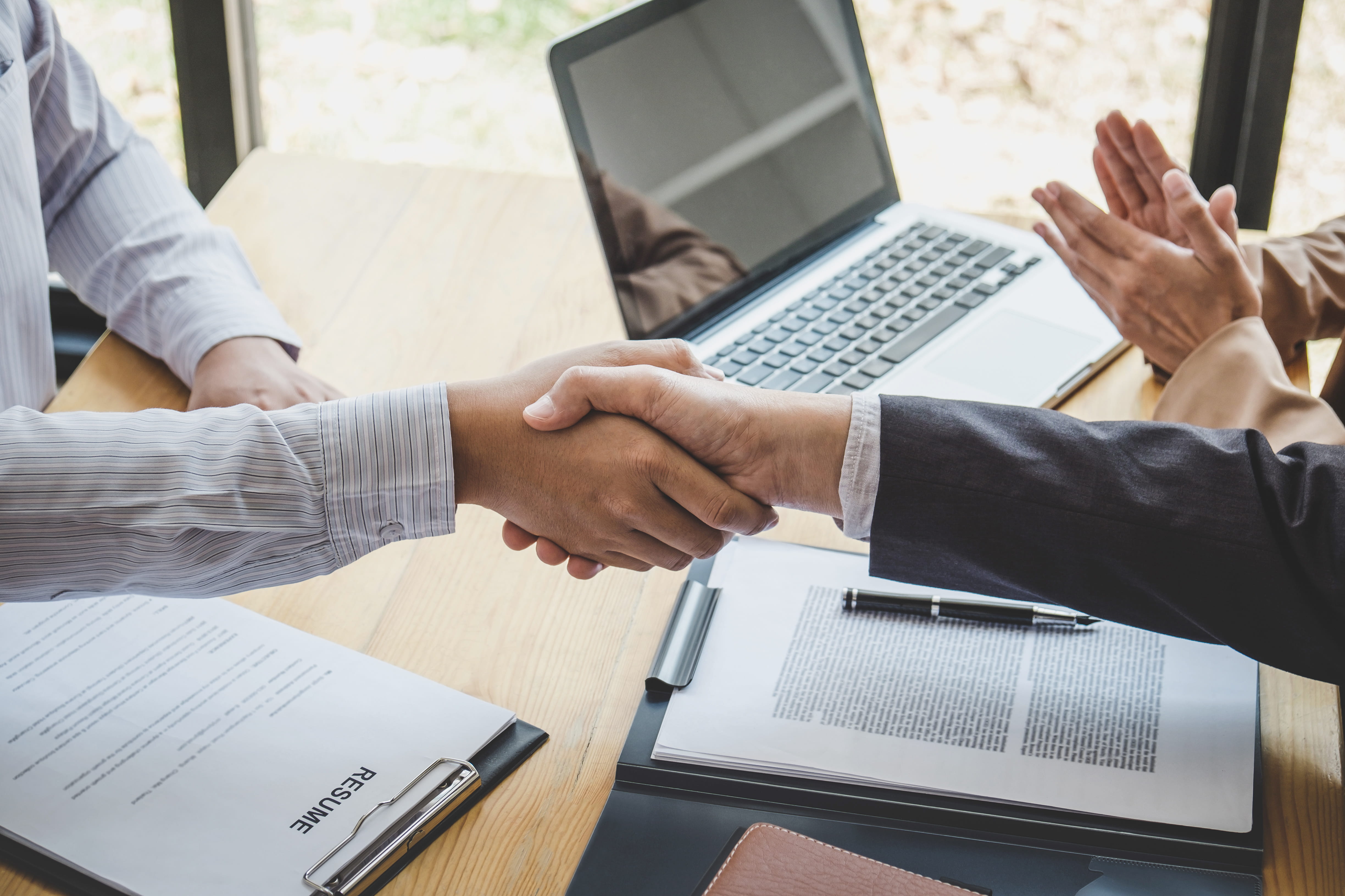 Close up of two business people shaking hands. There are papers and laptops in the background.