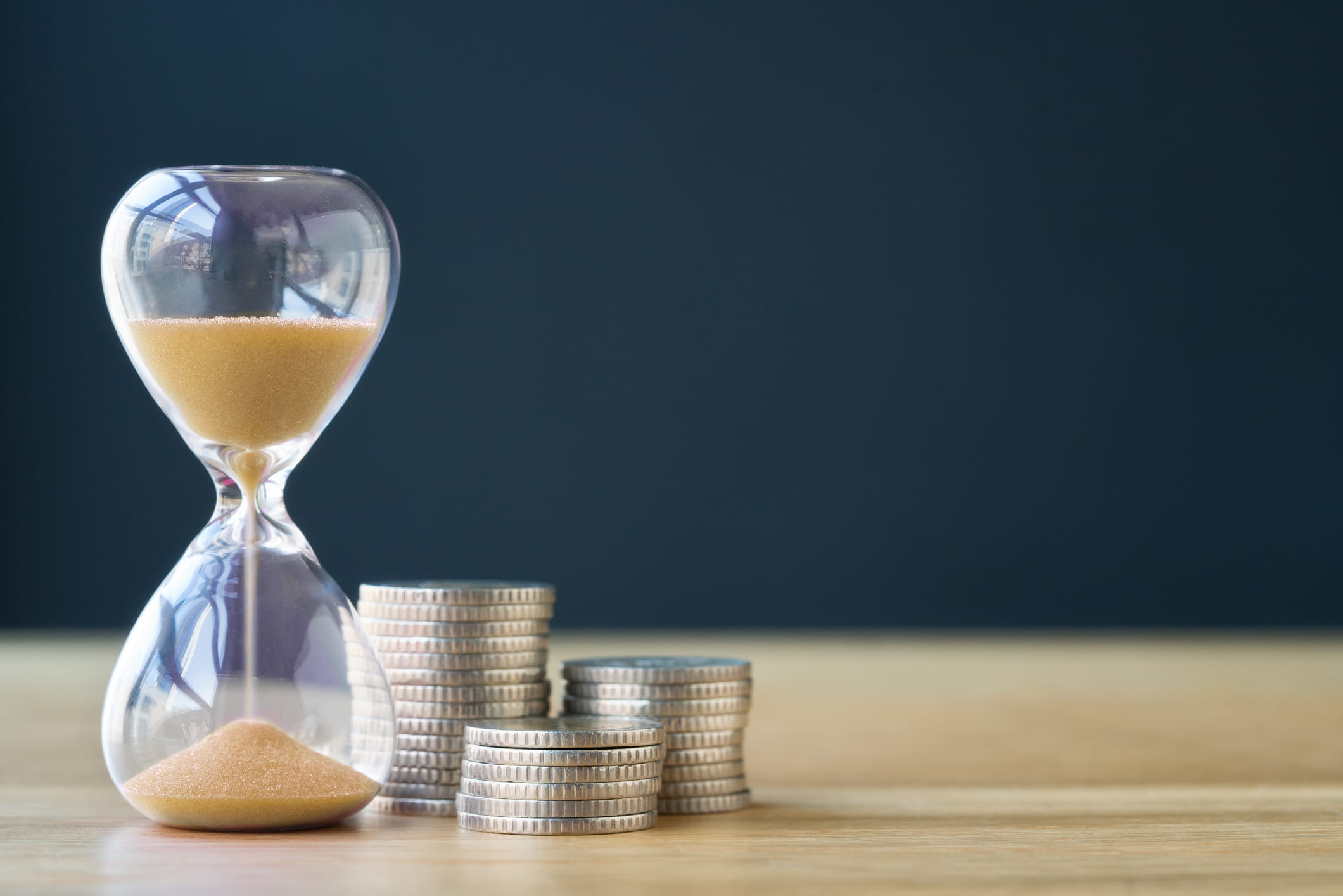 An hourglass with sand running through it stands next to several stacks of silver coins on a wooden surface against a dark blue background.