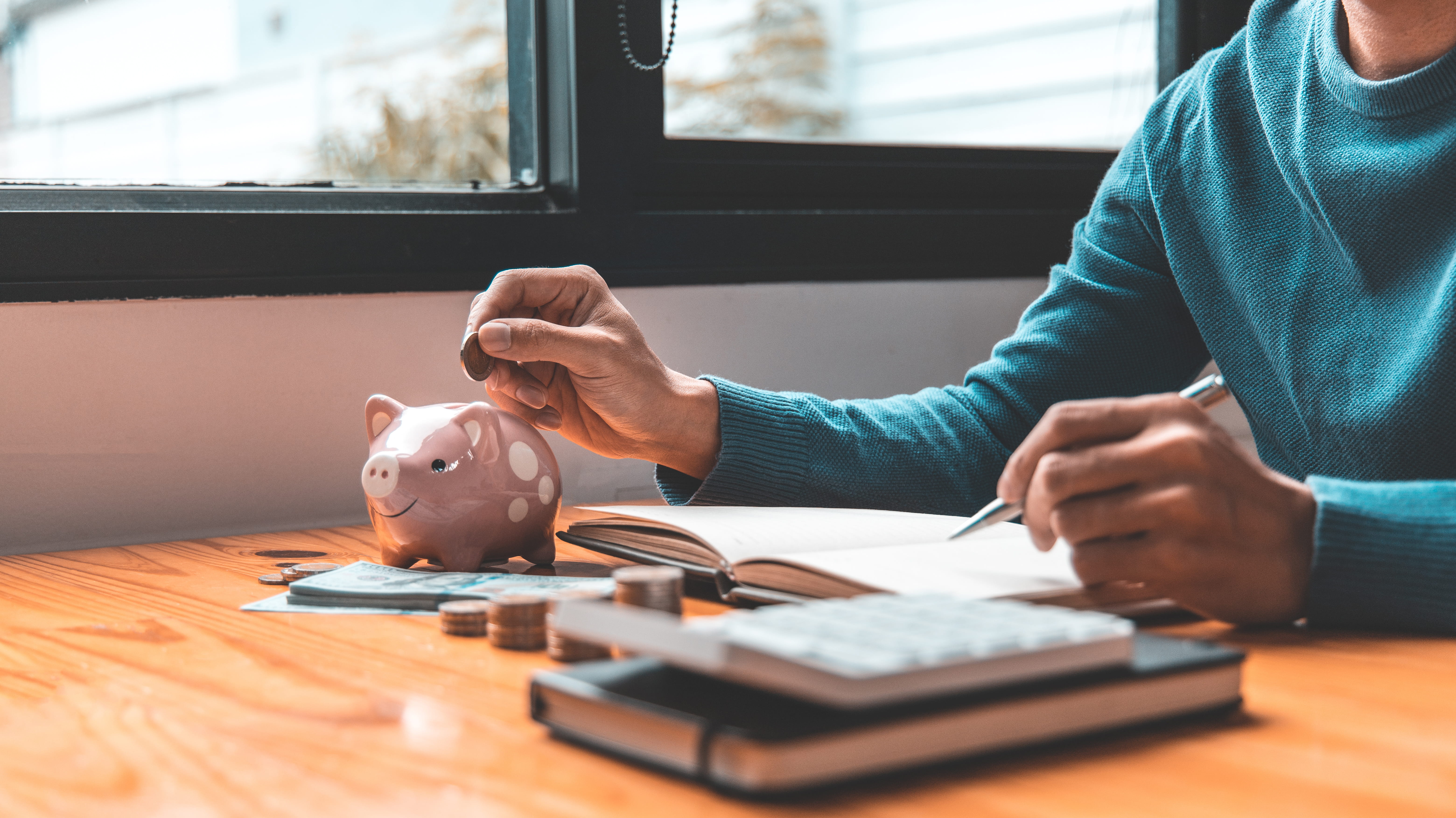 A person puts a coin into a pink piggy bank while writing in a notebook. Stacks of coins, cash, and a calculator are on the wooden desk.