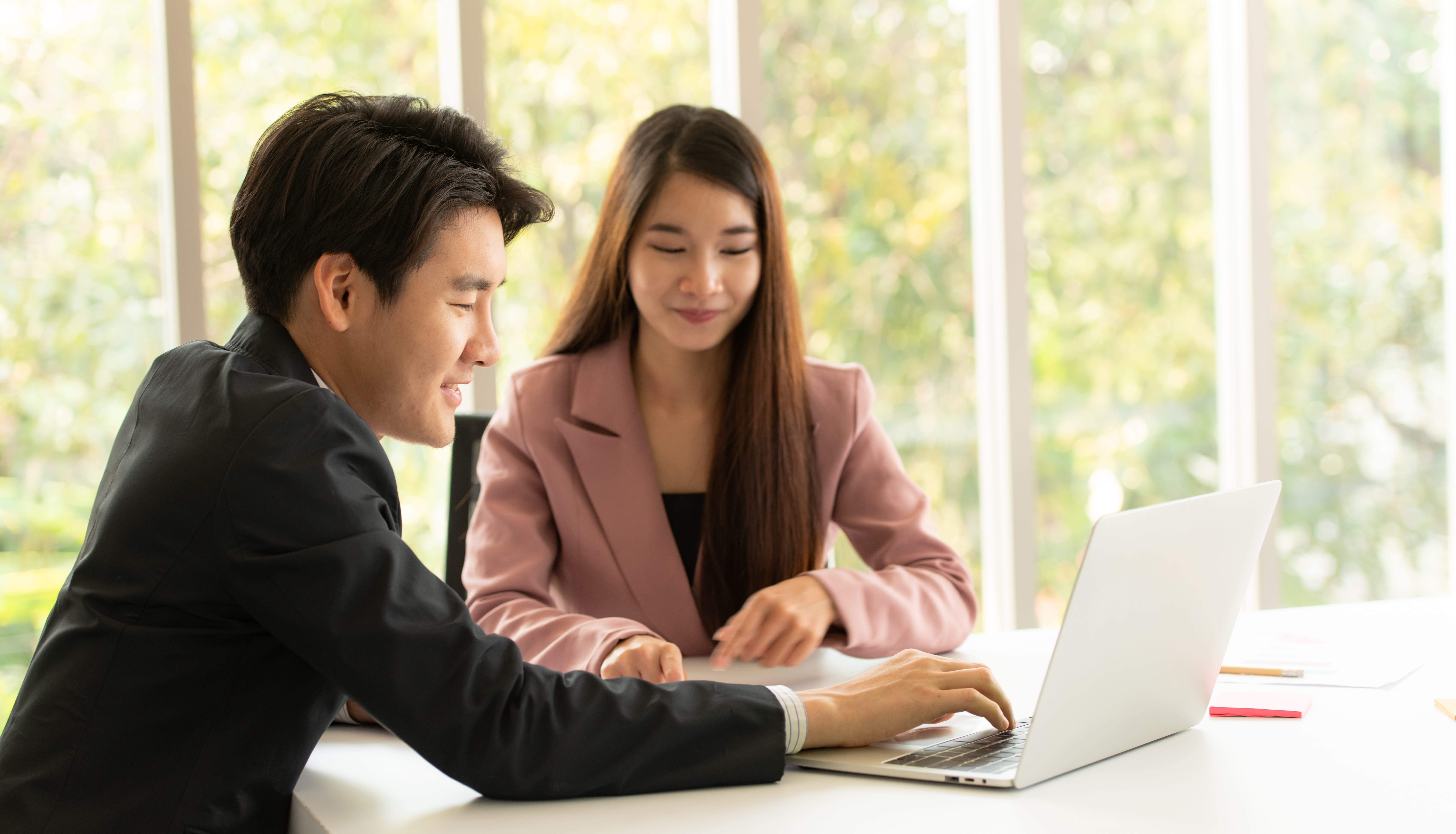 Two colleagues in professional attire sit at a white table in a bright office. A man in a suit uses a laptop while a woman in a pink blazer looks on. Large windows with trees are in the background.