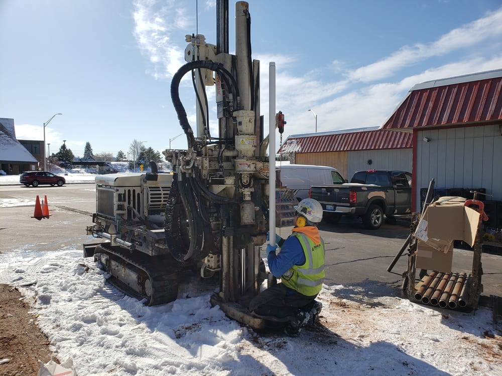 An EGLE RRD employee is kneeling on the ground near a large drilling machine; they are installing groundwater monitoring wells to check for groundwater contamination