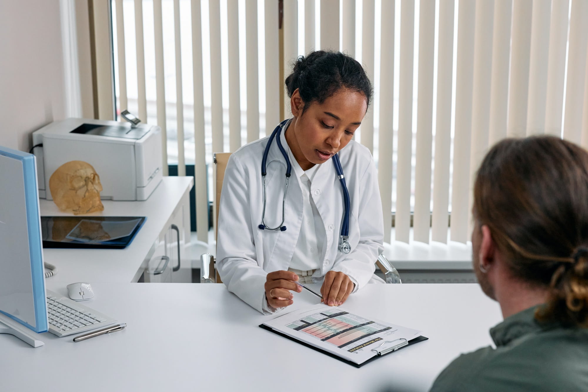 A female doctor siting at a desk in an office, going over a medical chart with a patient
