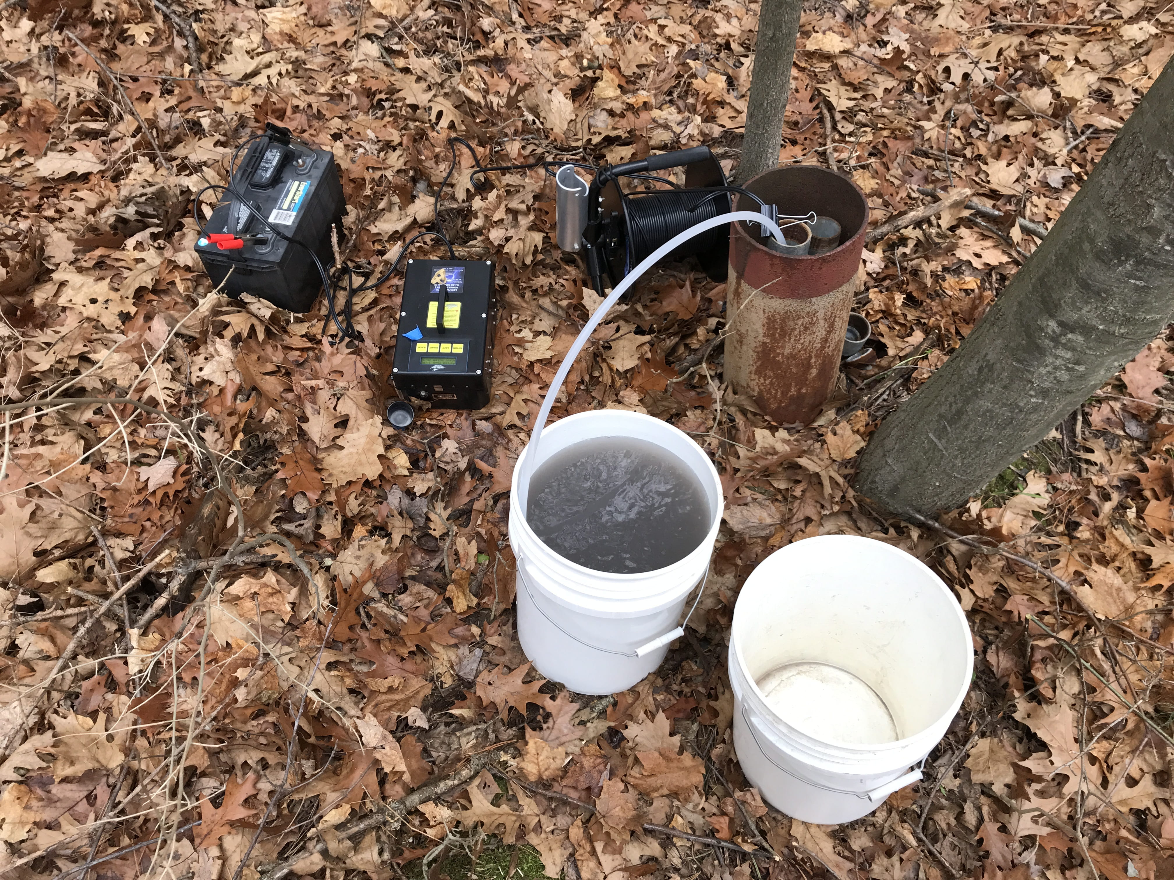 This is a typical submersible groundwater sampling set up.  A stainless steel PFAS free submersible pump is lowered into the well and into the groundwater table.  Tubing is connected to the pump which can be seen coming out of the well casing and into the bucket.  The submersible pump is connected to a Marine battery to provide power.  Groundwater can then be seen flowing through the tubing and into the 5 gallon bucket.  Three or more well volumes of water are purged from the monitoring well to ensure a representative groundwater sample is collected before filling laboratory bottles for analysis. 