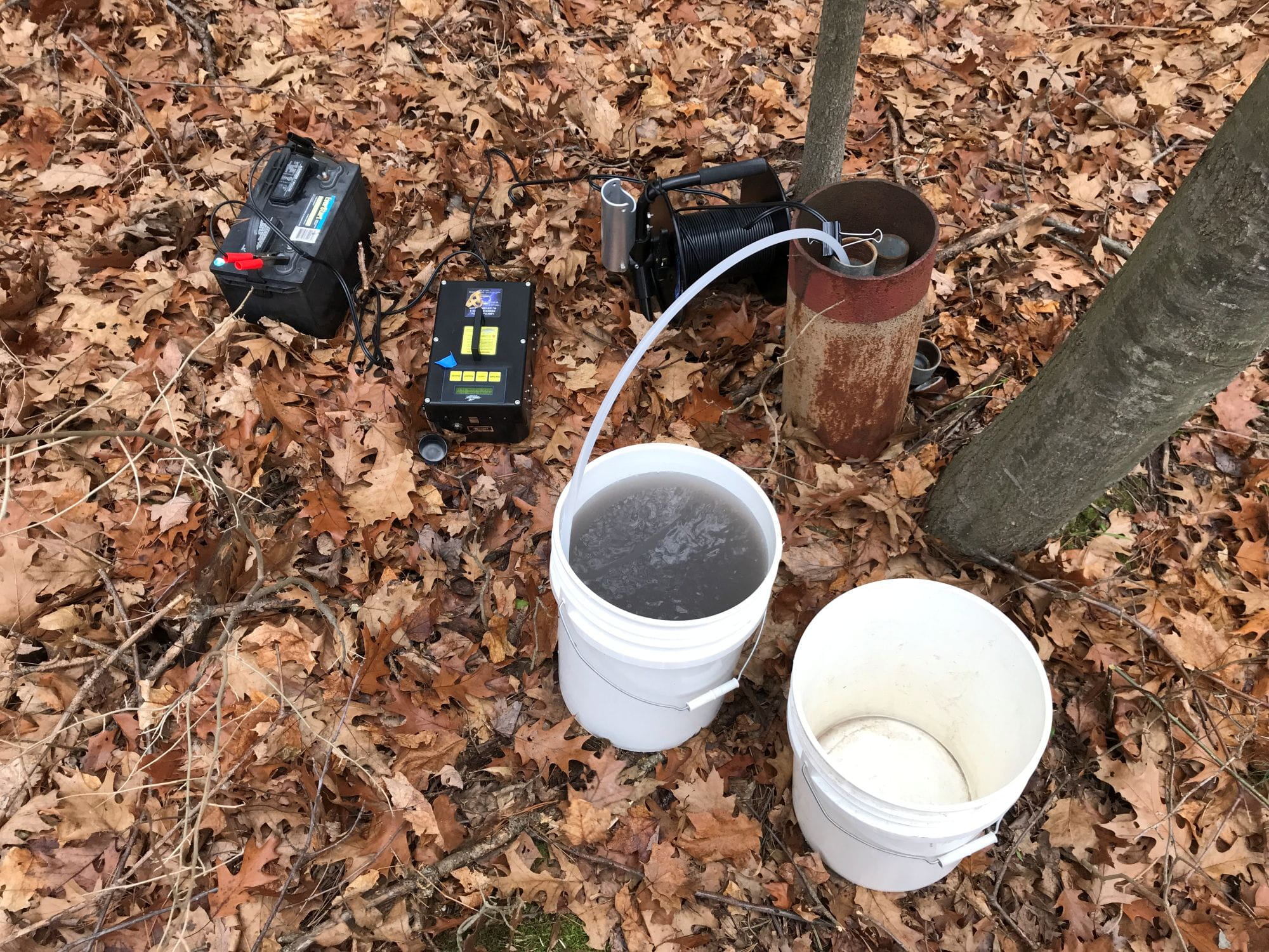 This is a typical submersible groundwater sampling set up.  A stainless steel PFAS free submersible pump is lowered into the well and into the groundwater table.  Tubing is connected to the pump which can be seen coming out of the well casing and into the bucket.  The submersible pump is connected to a Marine battery to provide power.  Groundwater can then be seen flowing through the tubing and into the 5 gallon bucket.  Three or more well volumes of water are purged from the monitoring well to ensure a representative groundwater sample is collected before filling laboratory bottles for analysis. 