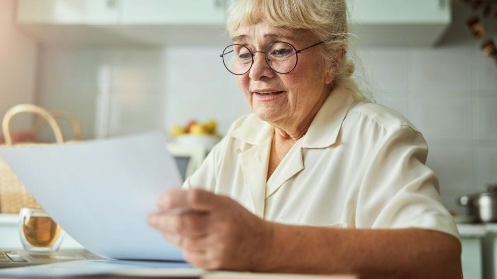An older woman wearing glasses and sitting at a kitchen table reading a sheet of paper