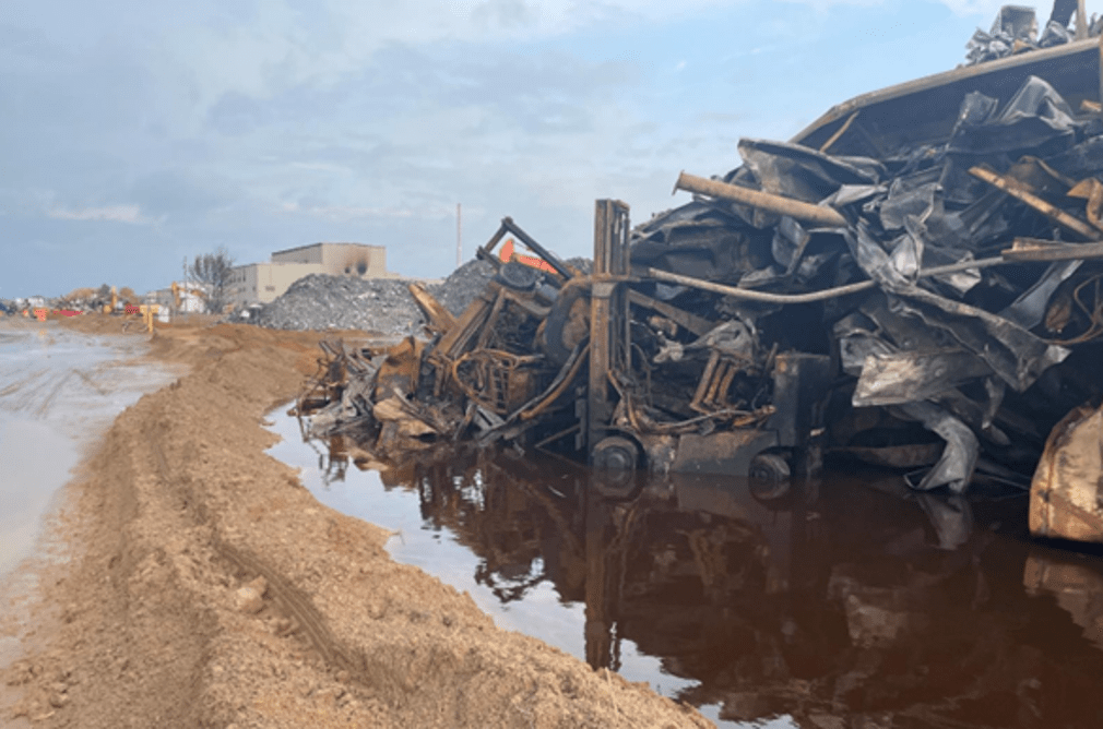A pile of twisted metal and other debris in a puddle of water, with the Menominee Warehouse in the background