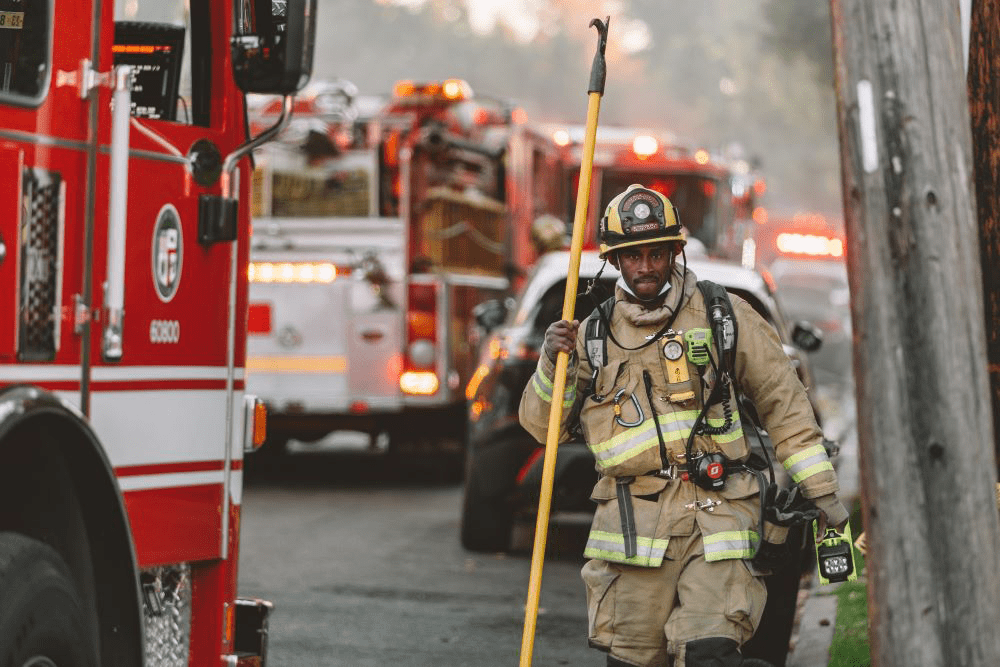 A determined-looking firefighter walks away from a cluster of firetrucks, loaded with equipment and holding a pole and electronic device