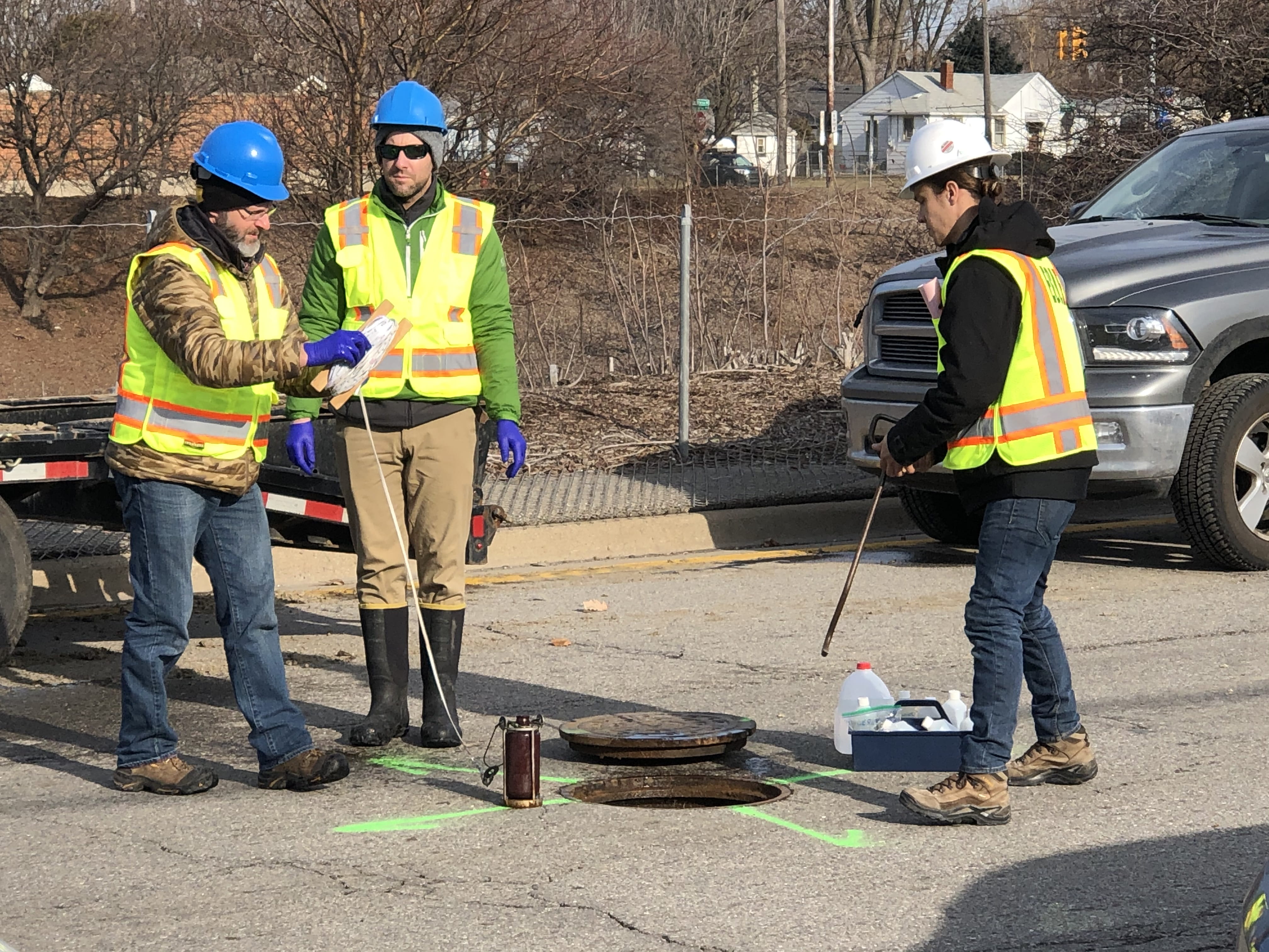 Three EGLE staff with sampling equipment outside at a roadway manhole. 