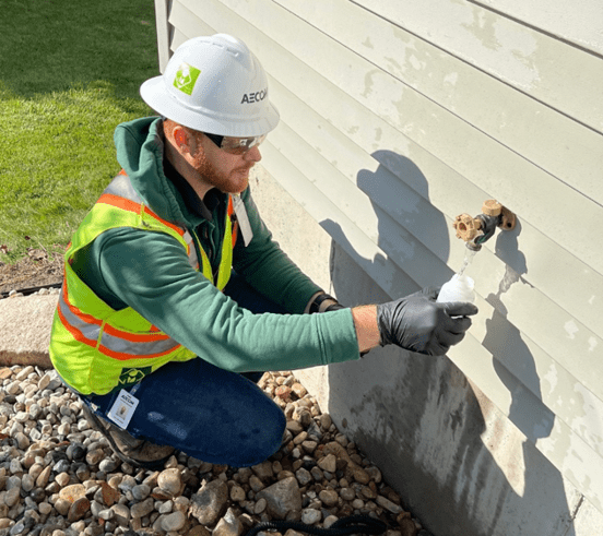 A person wearing a reflective vest and a hardhat kneeling outside a house at a faucet filling up a sample bottle