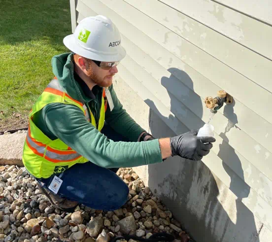 A person wearing a reflective vest and a hardhat kneeling outside a house at a faucet filling up a sample bottle