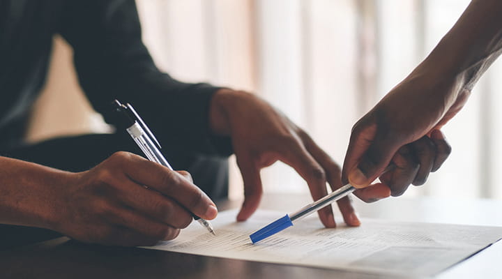 Two sets of black hands on a form. One set is filling out paperwork and the other is pointing to a section of the form. 
