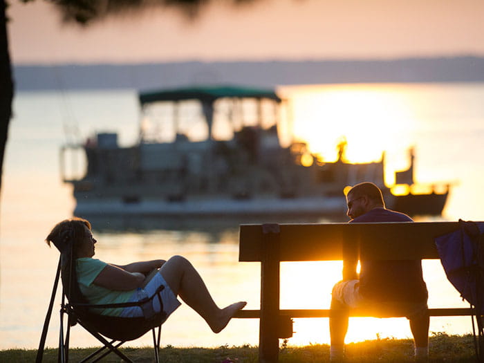 Two visitors sitting on bench and chair with boat in background