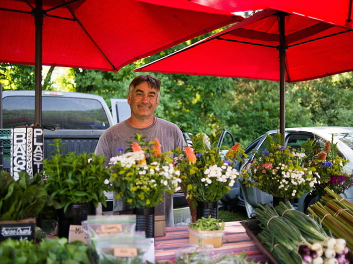 famers market vendor standing behind booth with veggies and flowers