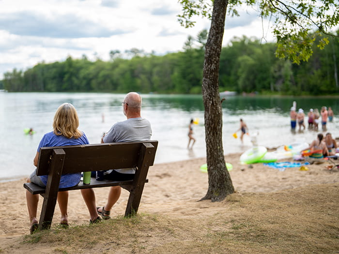 two visitors sitting on bench with beachgoers in background