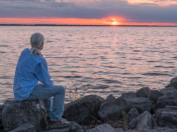 visitor sitting on rocks looking out to lake and sunset 