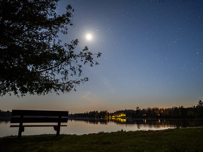 park bench set by water looking toward lit up campground in evening