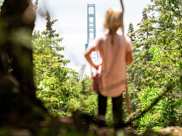 A young girl in the foreground looking through an opening in the trees toward the Mackinac Bridge