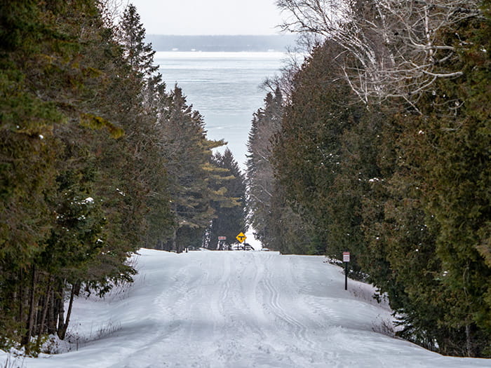 A snow-covered park road bordered by conifer trees overlooking the Straits of Mackinac