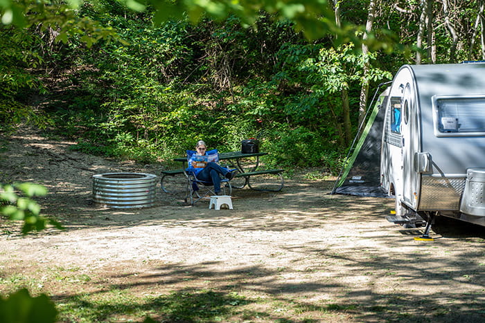 A seated person reading a book near a camping trailer on a wooded campsite