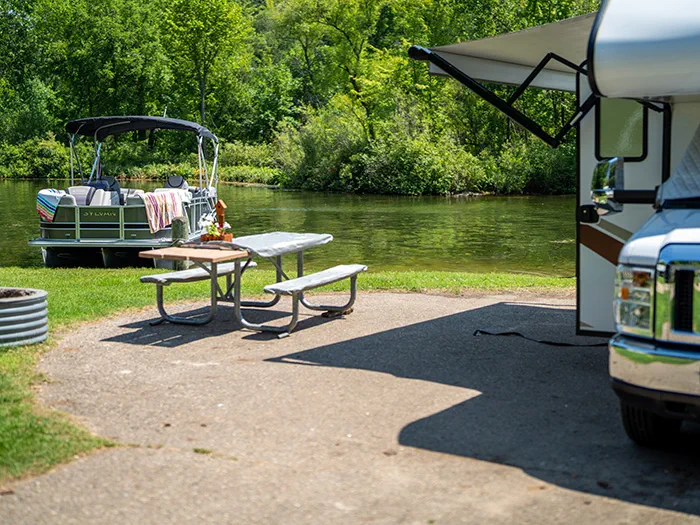 A modern campsite with fire ring, picnic table and camper parked on site with a boat docked in the background