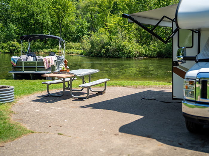 A modern campsite with fire ring, picnic table and camper parked on site with a boat docked in the background