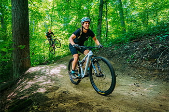 a mountain biker riding on trail with another biker getting air