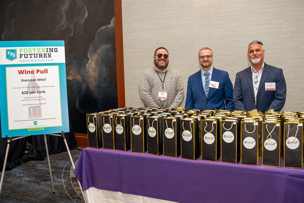 Three men standing behind a table filled with 50 bottles of wine, working the wine pull raffle
