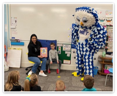 Mother and son reading to class. Owl mascot stands with them in front of the classroom