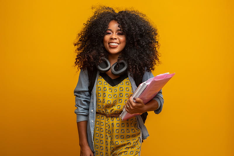 Young adult female student on campus with books in her arms, isolated on a yellow background