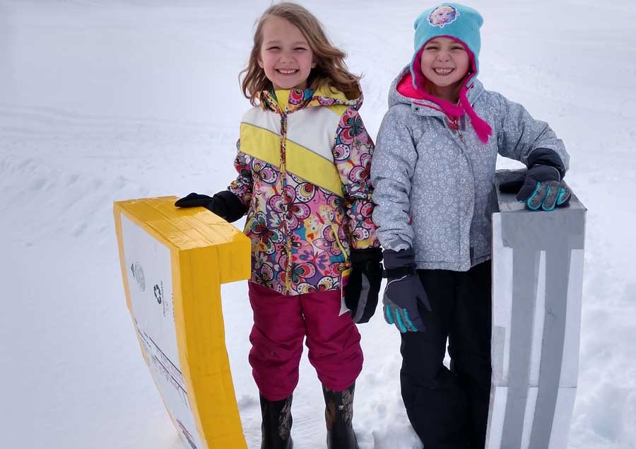 Two kids stand in the snow, smiling and holding their cardboard sleds