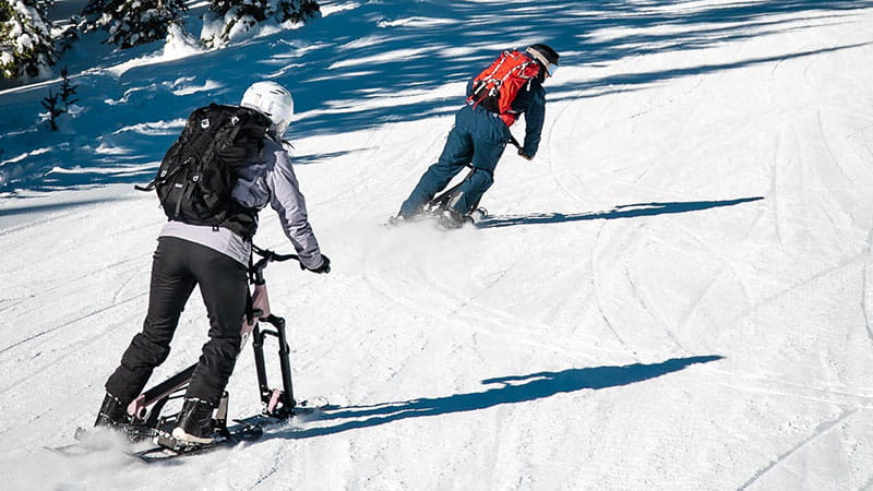 Two Sno-Go bikers ride down the slopes, dressed in warm winter clothing