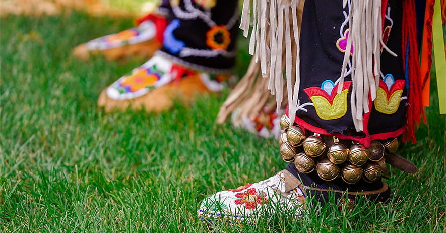 Image of two people standing in grass. The photo focuses on their lower legs and feet. They are dressed in native American dance and ceremonial attire. 