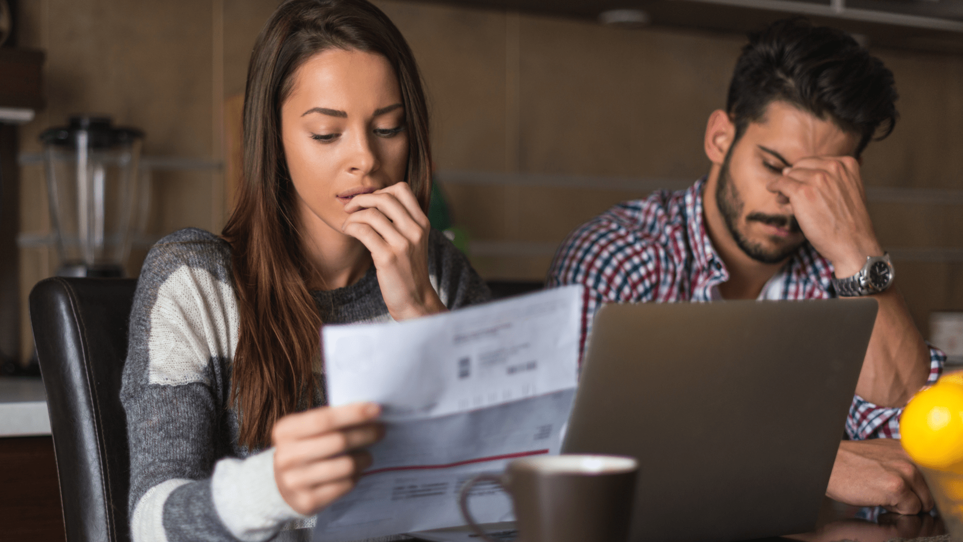 Photo of a woman looking at a piece of paper sitting down at a table. A man is sitting next to her with his head in his hands.