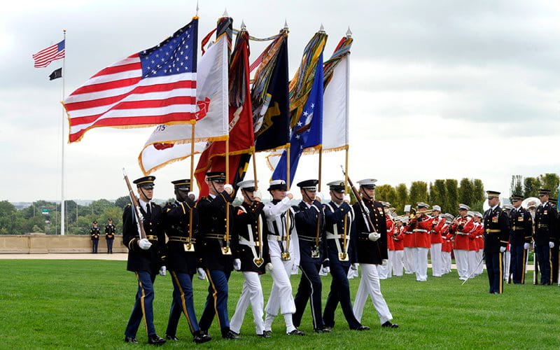 Photo of armed service members marching and carrying a flag.