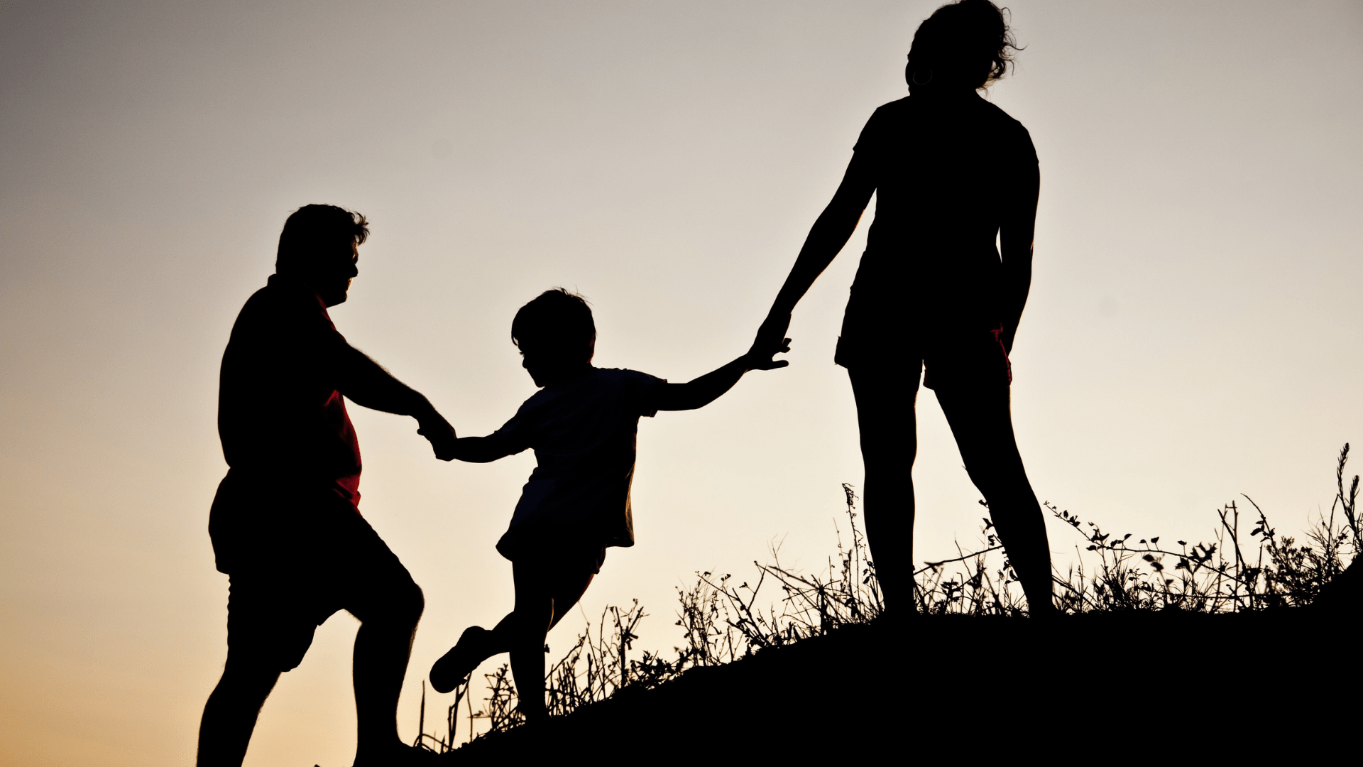 Silhouette  during sunset of two adults and a child outside climbing a hill.