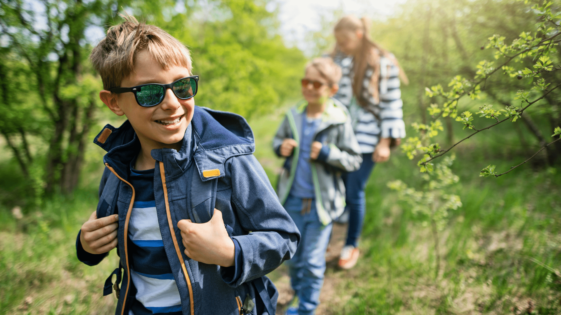 Image of three people hiking in the woods in springtime. They are smiling, wearing jackets, and wearing sunglasses. Trees are starting the bud in the background.