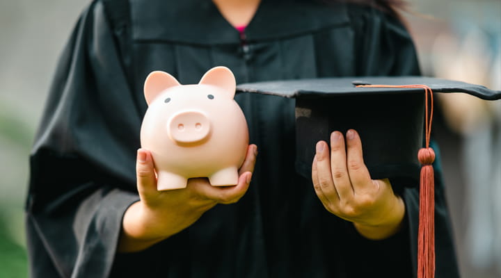 Woman in a graduation gown, holding a piggy bank and cap