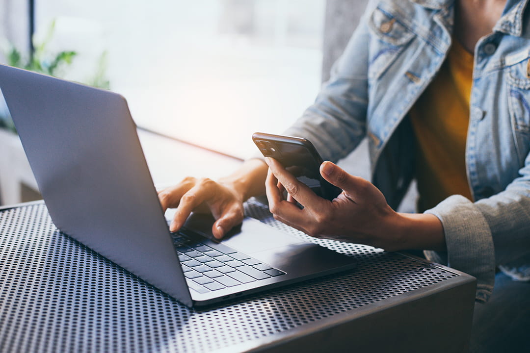 Person typing on a laptop computer while holding a mobile phone.