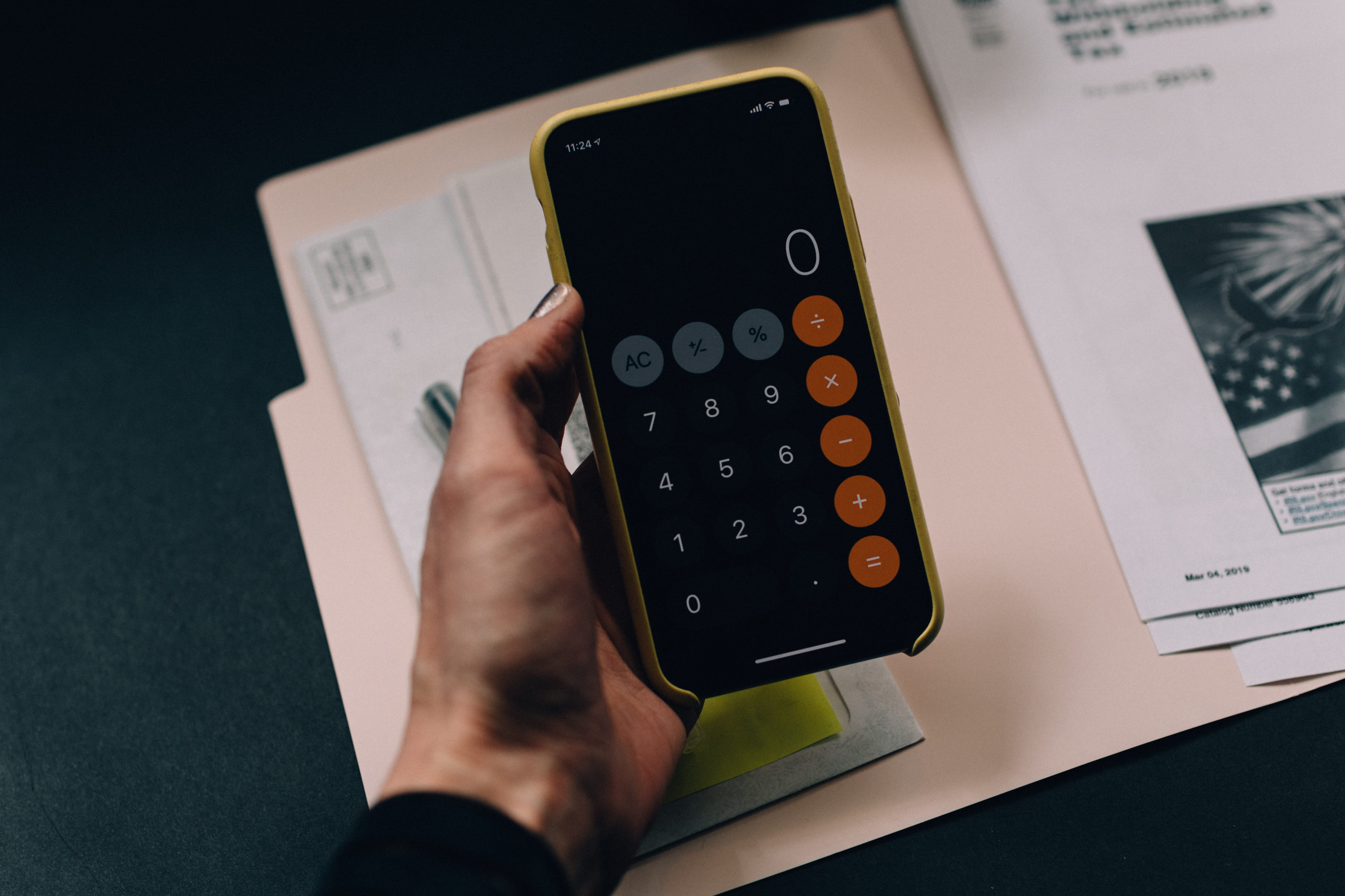 Hand holding a smartphone, using a calculator app, over a folder with tax documents.