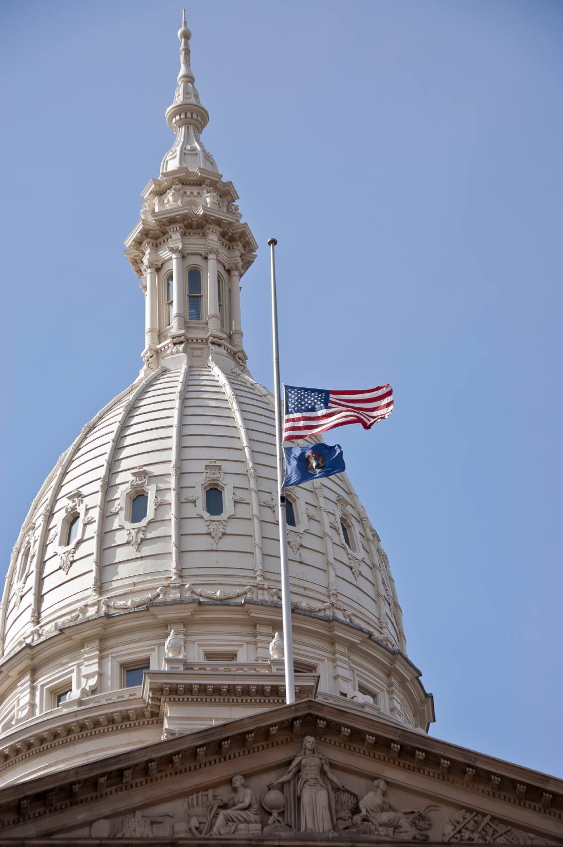 U.S. and Michigan flags flying in the wind at half staff atop a pole on the Michigan State Capitol building.