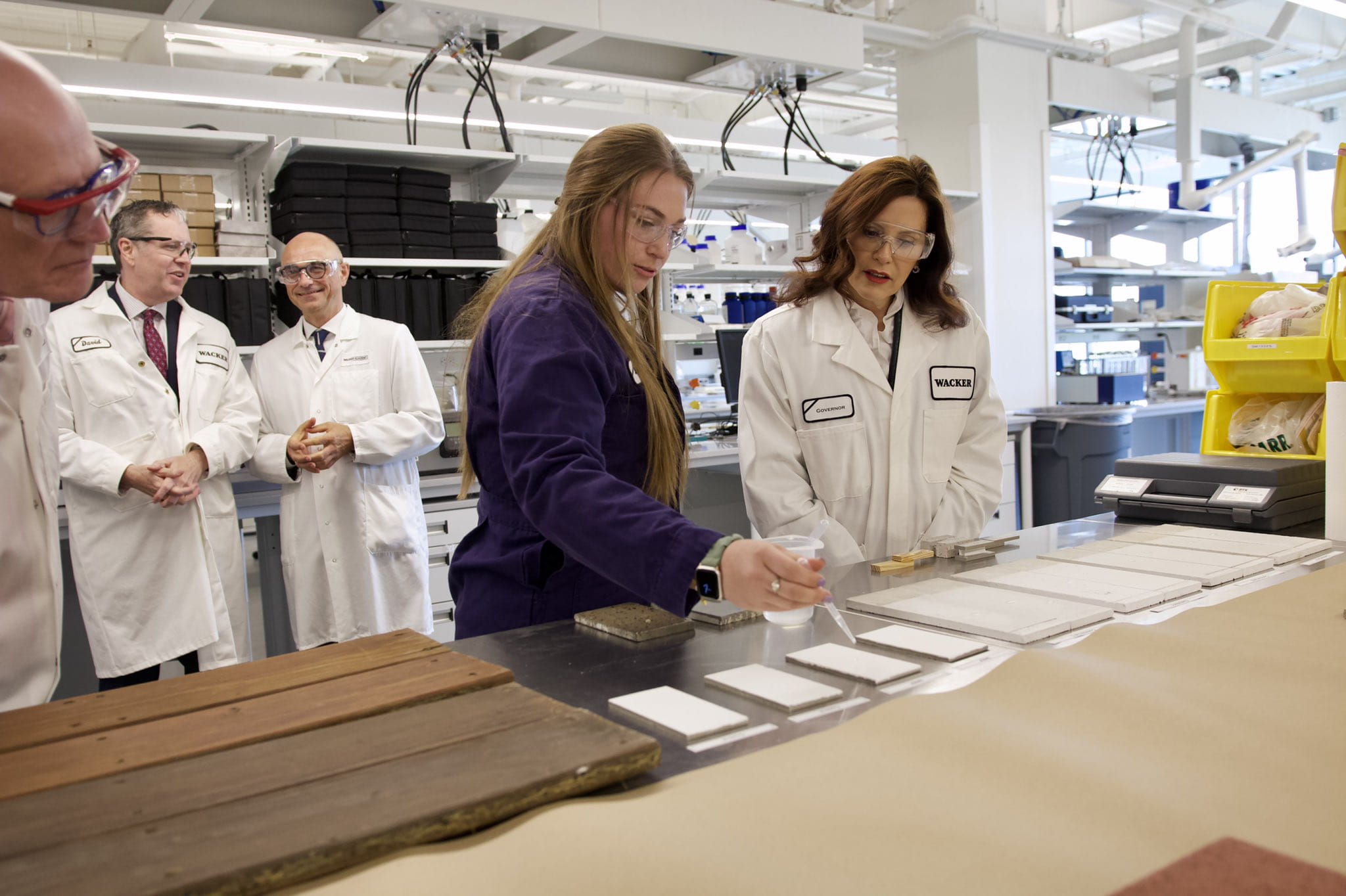 Governor Whitmer wears a lab coat and goggles as she watches a demonstration in a lab at Wacker Innovation Center.